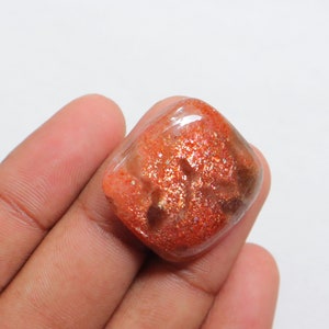 Pu&ograve; includere: Un cabochon di pietra di sole arancione, di forma quadrata, traslucido, con una finitura scintillante e iridescente. La pietra ha un colore marrone rossiccio con macchie dorate e argentate.