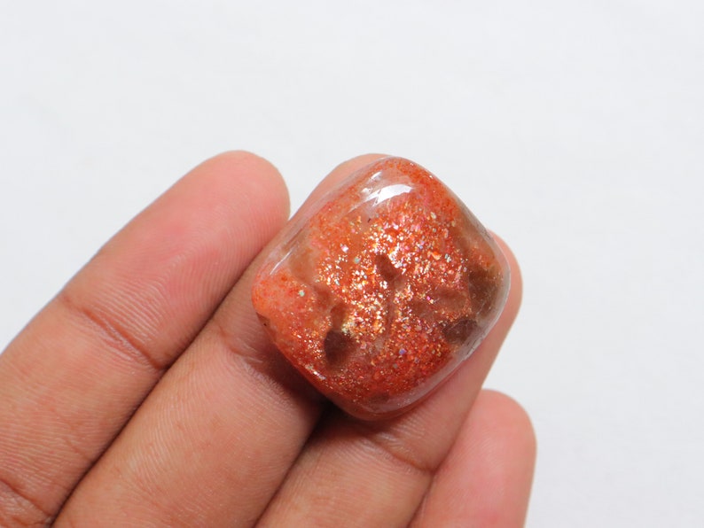Pu&ograve; includere: Un cabochon di pietra di sole arancione, di forma quadrata, traslucido, con una finitura scintillante e iridescente. La pietra ha un colore marrone rossiccio con macchie dorate e argentate.