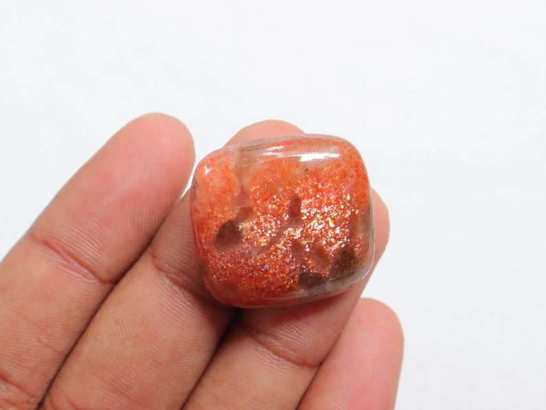 Pu&ograve; includere: Un cabochon di pietra di sole di forma quadrata, di colore arancione con una finitura scintillante e iridescente. La pietra ha un aspetto traslucido con una miscela di tonalit&agrave; arancioni, marroni e dorate.
