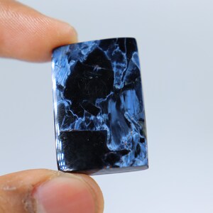 May include: A rectangular gemstone with a polished surface. The stone features a striking pattern of deep blue and black, creating a marbled effect. The gemstone is held between two fingers against a white background.