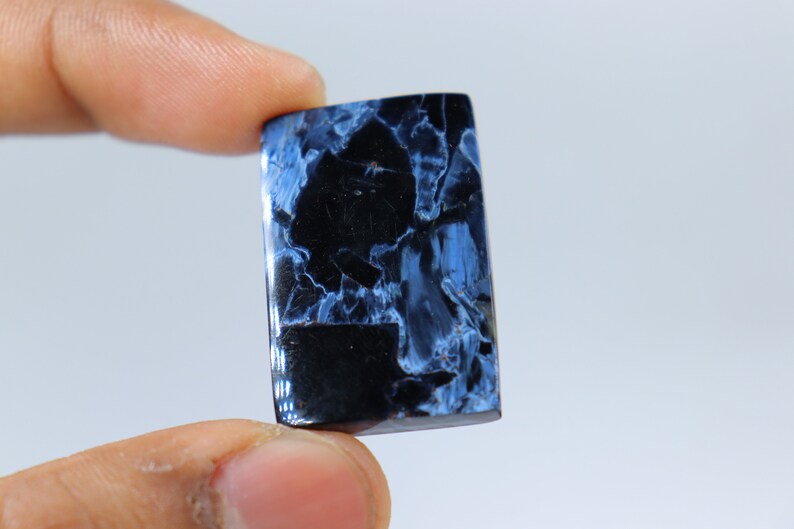 May include: A rectangular gemstone with a polished surface. The stone features a striking pattern of deep blue and black, creating a marbled effect. The gemstone is held between two fingers against a white background.