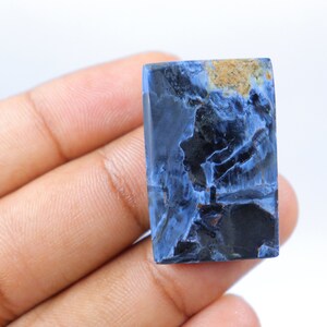 May include: A rectangular gemstone with a striking pattern of deep blue, black, and tan hues. The polished surface reveals a unique, marbled appearance, showcasing natural mineral formations. The stone is held in a hand against a white background.