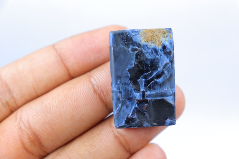May include: A rectangular gemstone with a striking pattern of deep blue, black, and tan hues. The polished surface reveals a unique, marbled appearance, showcasing natural mineral formations. The stone is held in a hand against a white background.