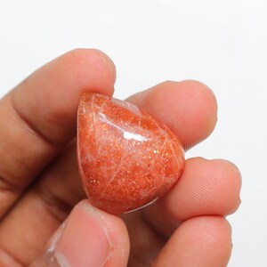 Pu&ograve; includere: Un cabochon di pietra di sole arancione lucido e liscio, con una tonalit&agrave; arancio rossastra e un leggero luccichio. La pietra &egrave; tenuta in una mano.