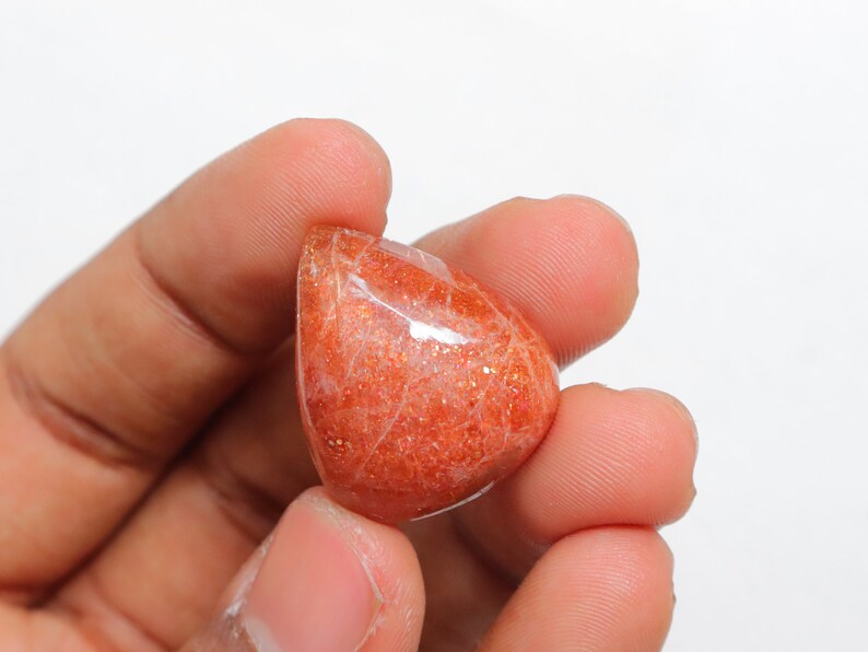 Pu&ograve; includere: Un cabochon di pietra di sole arancione lucido e liscio, con una tonalit&agrave; arancio rossastra e un leggero luccichio. La pietra &egrave; tenuta in una mano.