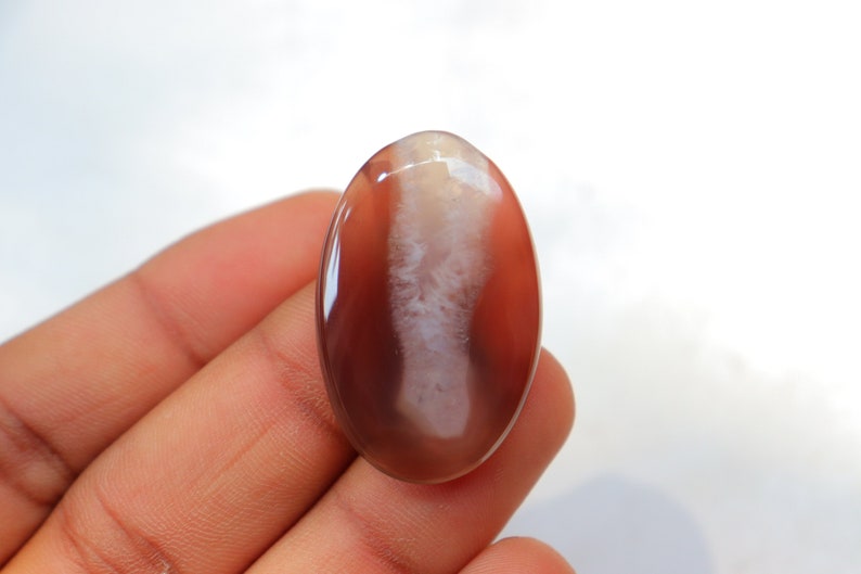 May include: A smooth, oval-shaped agate cabochon with a white and brown pattern. The stone is translucent and has a glossy finish.