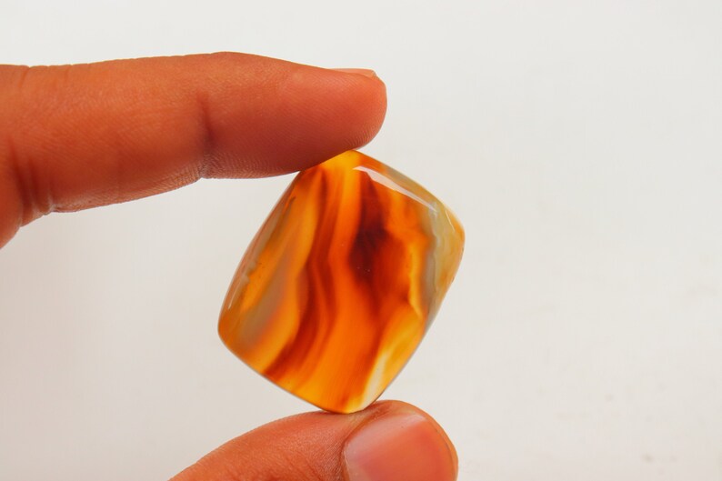 Op de afbeelding: Een gladde, gepolijste, oranje agaat cabochon met witte en bruine banden. De steen is rechthoekig van vorm en heeft een glanzende afwerking.