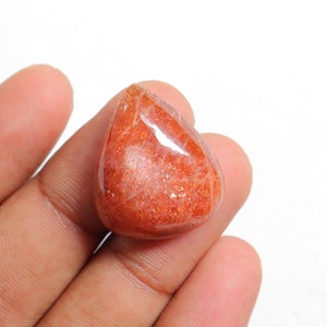 Pu&ograve; includere: Un cabochon di pietra di sole arancione a forma di lacrima con una finitura scintillante e iridescente. La pietra &egrave; tenuta in una mano, mostrando le sue dimensioni e la sua forma.