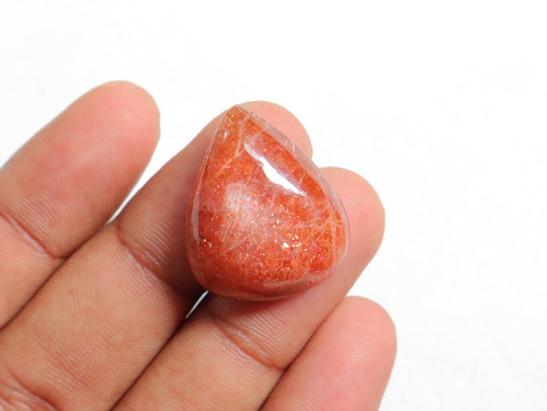 Pu&ograve; includere: Un cabochon di pietra di sole arancione a forma di lacrima con una finitura scintillante e iridescente. La pietra &egrave; tenuta in una mano, mostrando le sue dimensioni e la sua forma.