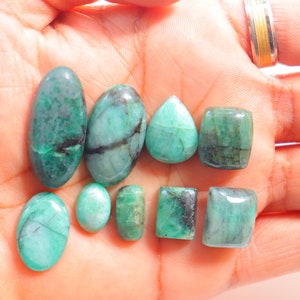May include: A collection of eight emerald green gemstones of various shapes and sizes, including oval, pear, and rectangular cuts. The gemstones are displayed on a person's hand.