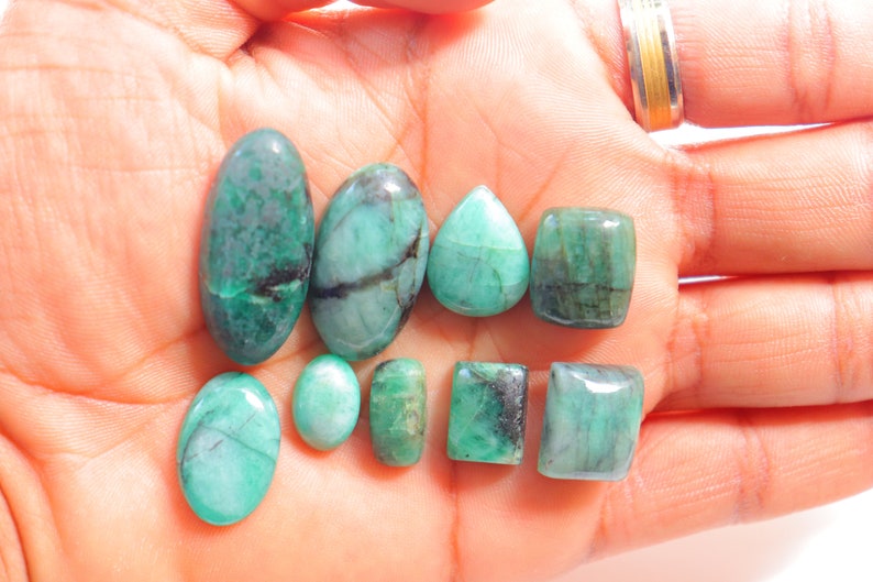 May include: A collection of eight emerald green gemstones of various shapes and sizes, including oval, pear, and rectangular cuts. The gemstones are displayed on a person's hand.