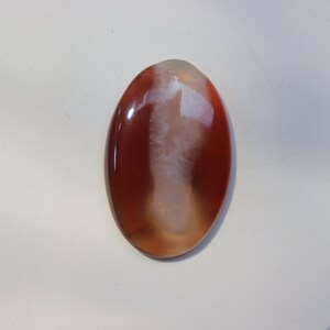 May include: A smooth, oval-shaped agate cabochon with a white and brown pattern. The stone has a glossy finish.