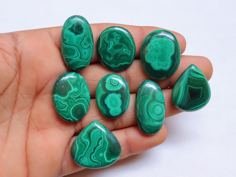 May include: A collection of eight green malachite cabochons, each with a unique pattern of swirling veins and a polished, smooth surface.