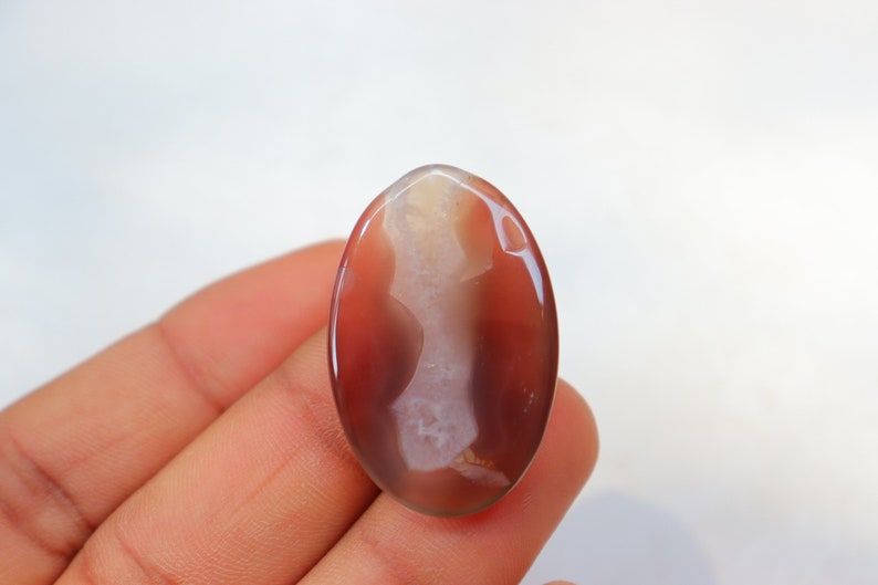 May include: A smooth, oval-shaped agate gemstone with a white and brown pattern. The stone is translucent and has a glossy finish.