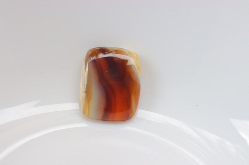 Op de afbeelding: Een gladde, gepolijste agaat cabochon met bruine, witte en oranje banden. De steen is rechthoekig en heeft een glanzende afwerking.