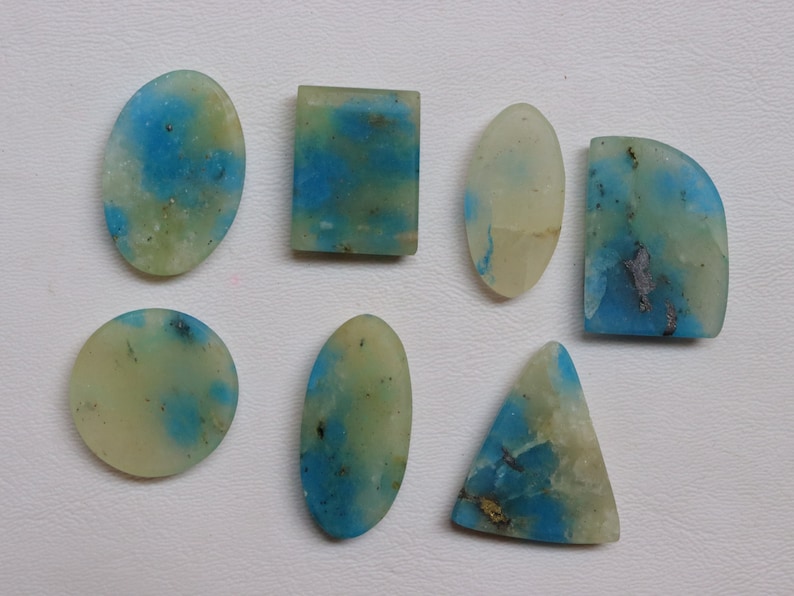 May include: Seven cabochon gemstones in various shapes and sizes, including oval, round, rectangle, and triangle. The stones are a pale yellow-green color with blue and gray inclusions.