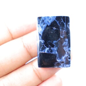 May include: A rectangular gemstone with a polished surface. The stone features a dark blue and black color scheme with white veining throughout. The gemstone is held in a hand against a white background.