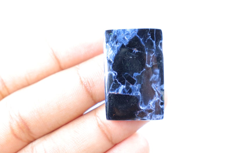 May include: A rectangular gemstone with a polished surface. The stone features a dark blue and black color scheme with white veining throughout. The gemstone is held in a hand against a white background.