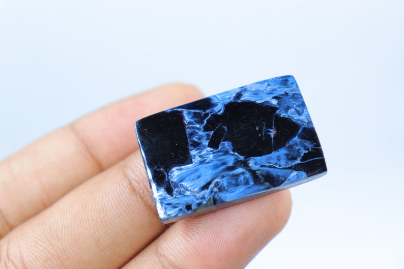 May include: A rectangular gemstone with a polished surface. The stone features a striking pattern of deep blue and black swirls and veins. The gemstone is held in a hand against a white background.