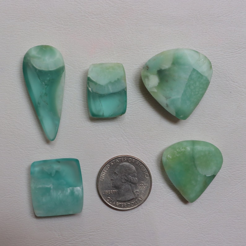 May include: Five green and white gemstone cabochons of various shapes and sizes, including a teardrop, rectangle, triangle, and two heart shapes. The gemstones are arranged on a white surface with a US quarter dollar coin for scale.