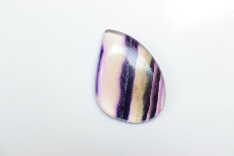 May include: A teardrop-shaped cabochon gemstone with a white, purple, and yellow striped pattern.