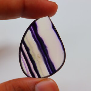 May include: A teardrop-shaped, translucent purple and white fluorite gemstone with a banded pattern.