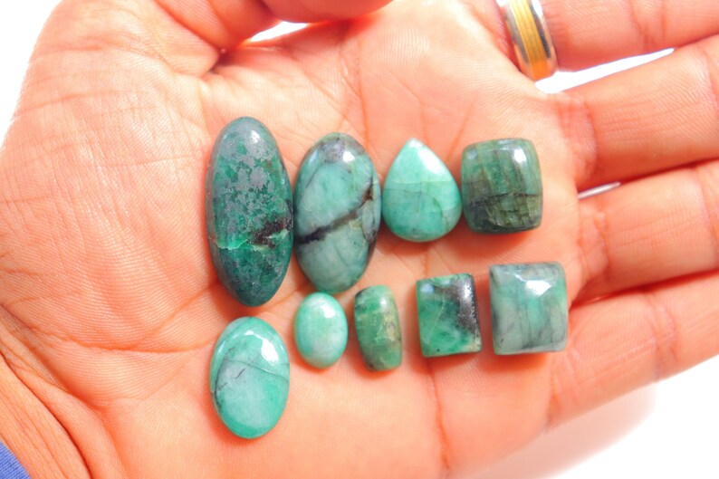 May include: A collection of eight emerald green gemstones of various shapes and sizes, including oval, pear, and rectangular cuts. The gemstones are displayed on a hand.