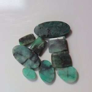 May include: A collection of eight green emerald gemstones in various shapes and sizes. The stones are a dark green colour with black and white inclusions.