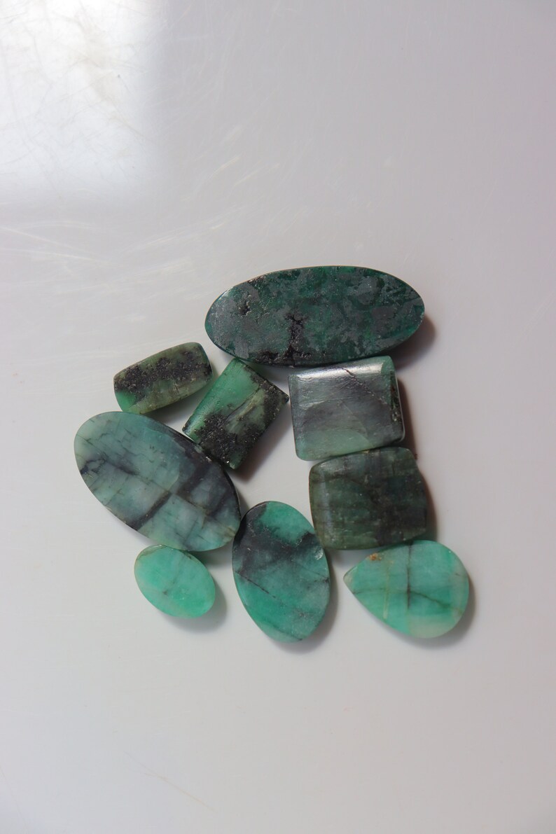 May include: A collection of eight green emerald gemstones in various shapes and sizes. The stones are a dark green colour with black and white inclusions.