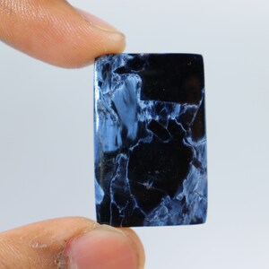 May include: A rectangular gemstone with a polished surface. The stone features a striking pattern of deep black and light blue veining, creating a marbled effect. The gemstone is held between two fingers against a plain white background.