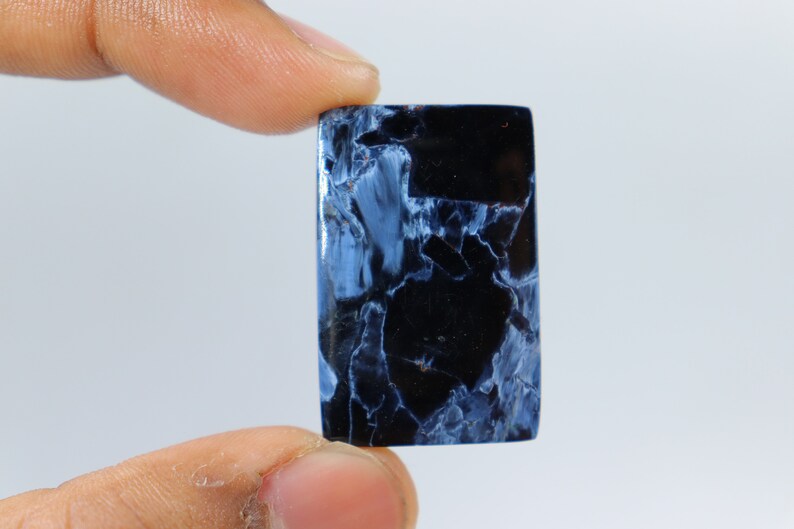 May include: A rectangular gemstone with a polished surface. The stone features a striking pattern of deep black and light blue veining, creating a marbled effect. The gemstone is held between two fingers against a plain white background.