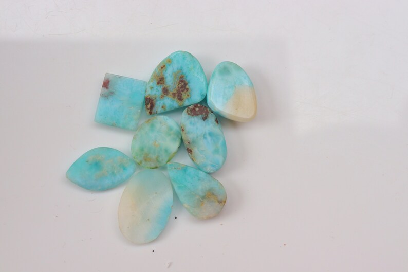 May include: A collection of eight turquoise cabochons, some with brown inclusions, in various shapes and sizes. The cabochons are arranged on a white surface.