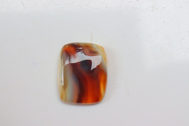 Op de afbeelding: Een gladde, gepolijste agaat cabochon met een rechthoekige vorm. De steen heeft een mix van witte, bruine en oranje kleuren in een draaiende patroon.