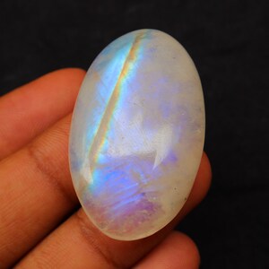 K&ouml;nnte beinhalten: Ein wei&szlig;er ovaler Mondstein-Cabochon mit schillernden blauen und violetten Blitzen. Der Stein wird in einer Hand gehalten.