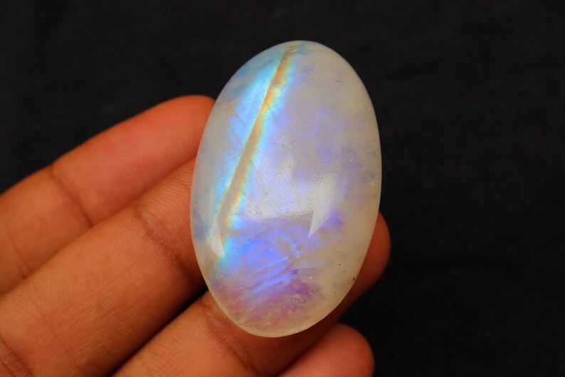 K&ouml;nnte beinhalten: Ein wei&szlig;er ovaler Mondstein-Cabochon mit schillernden blauen und violetten Blitzen. Der Stein wird in einer Hand gehalten.