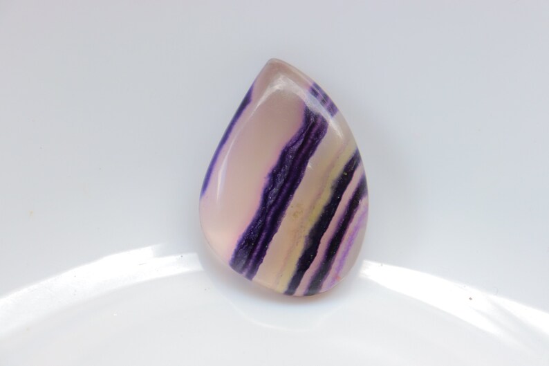 May include: A teardrop-shaped cabochon gemstone with purple and white stripes. The stone is translucent and has a smooth, polished surface.