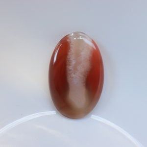 May include: A smooth, oval-shaped agate cabochon with a white and brown pattern. The agate is translucent and has a glossy finish.