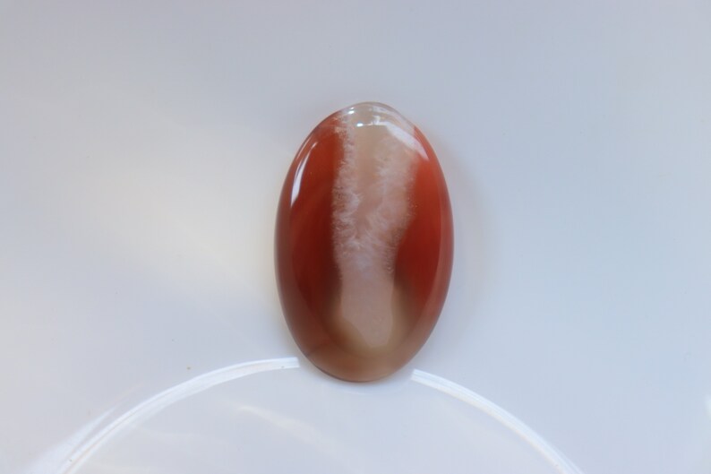 May include: A smooth, oval-shaped agate cabochon with a white and brown pattern. The agate is translucent and has a glossy finish.