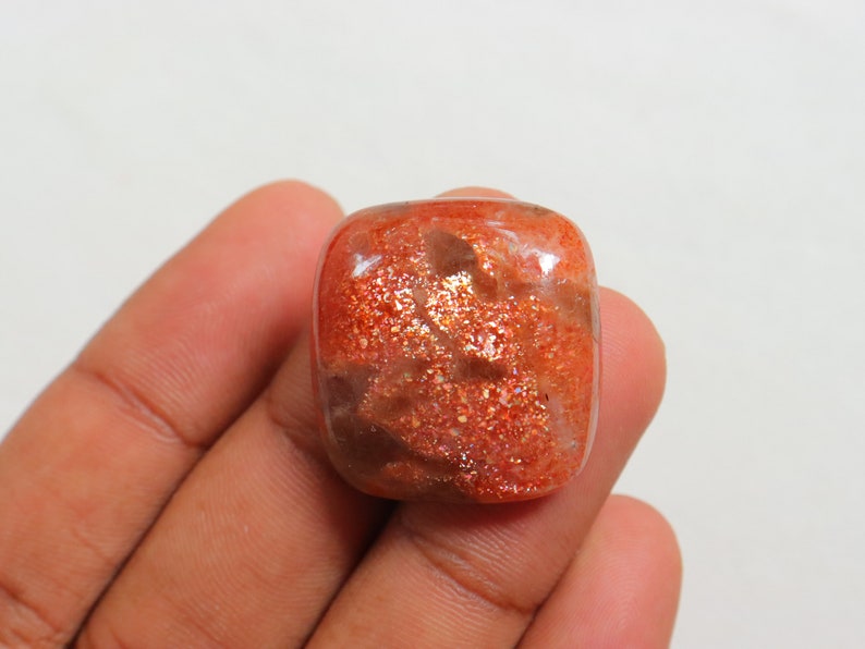 Pu&ograve; includere: Un grande cabochon di pietra di sole rettangolare di colore arancione con una finitura scintillante e iridescente. La pietra presenta una miscela di colori arancio, marrone e oro.