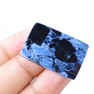 May include: A rectangular gemstone with a polished surface. The stone features a striking pattern of deep blue and black hues, creating a marbled effect. The gemstone is held against a white background.