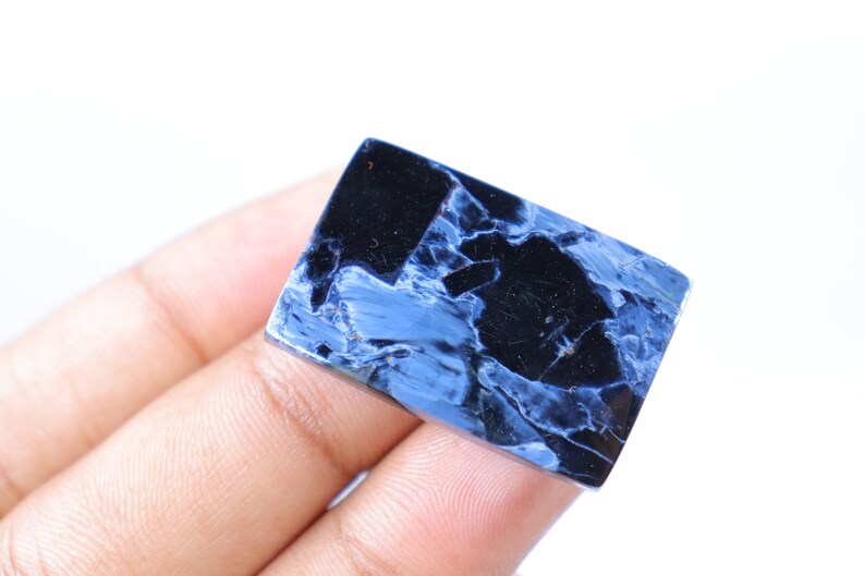May include: A rectangular gemstone with a polished surface. The stone features a striking pattern of deep blue and black hues, creating a marbled effect. The gemstone is held against a white background.