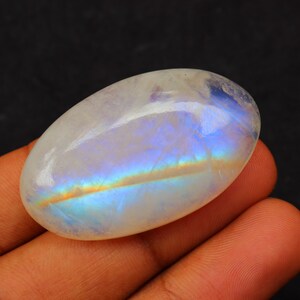 K&ouml;nnte beinhalten: Ein wei&szlig;er ovaler Mondstein-Cabochon mit irisierenden blauen und goldenen Schimmern. Der Stein ist glatt und poliert.