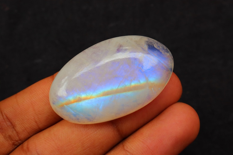 K&ouml;nnte beinhalten: Ein wei&szlig;er ovaler Mondstein-Cabochon mit irisierenden blauen und goldenen Schimmern. Der Stein ist glatt und poliert.