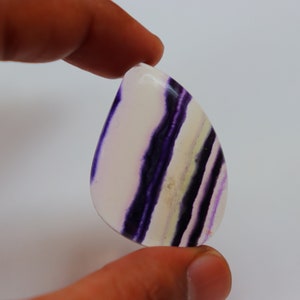 May include: A teardrop-shaped cabochon of purple and white fluorite with a banded pattern. The stone is translucent and has a smooth, polished surface.