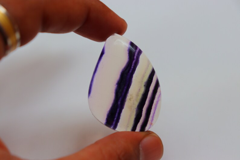 May include: A teardrop-shaped cabochon of purple and white fluorite with a banded pattern. The stone is translucent and has a smooth, polished surface.