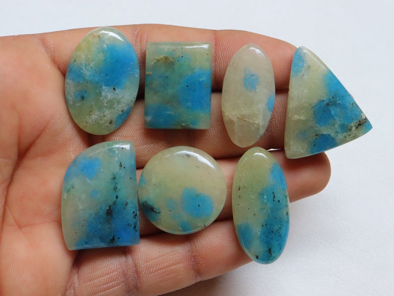 May include: Seven cabochon gemstones with a white and blue marbled pattern. The stones are various shapes and sizes, including oval, round, and rectangular.