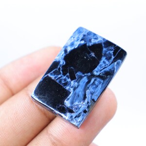 May include: A rectangular gemstone with a polished surface. The stone features a striking pattern of deep blue and black veining, creating a marbled effect. The gemstone is held in a hand against a white background.