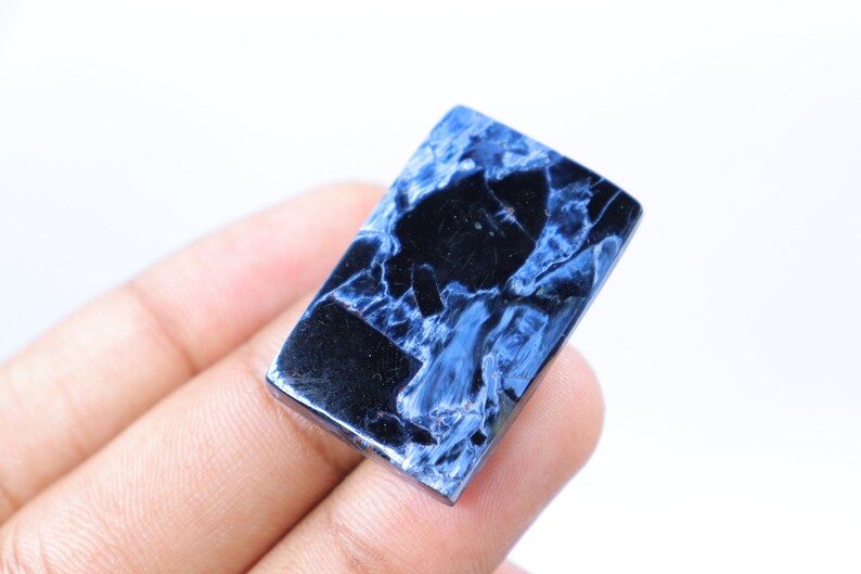 May include: A rectangular gemstone with a polished surface. The stone features a striking pattern of deep blue and black veining, creating a marbled effect. The gemstone is held in a hand against a white background.