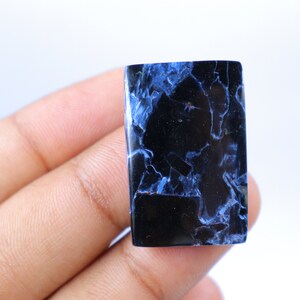 May include: A rectangular gemstone with a polished surface. The stone is primarily black with striking blue veining and patterns throughout. The gemstone is held in a hand against a white background.