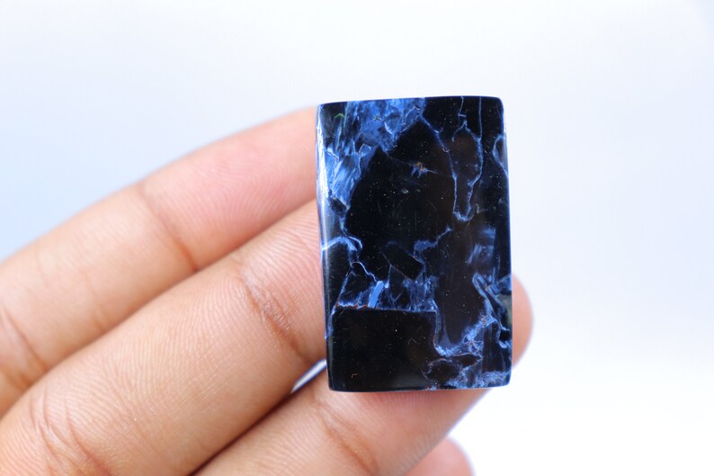 May include: A rectangular gemstone with a polished surface. The stone is primarily black with striking blue veining and patterns throughout. The gemstone is held in a hand against a white background.